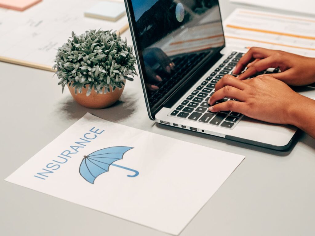 Hands typing on a laptop at a desk with an insurance paper and plant, suggesting a work environment.